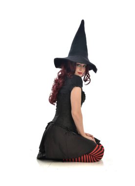 tam uzunlukta portresi kırmızı saçlı kız kara cadı kostüm ve sivri şapka giyiyor. beyaz stüdyo arka plan üzerinde izole oturmuş poz.