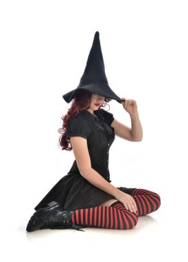 tam uzunlukta portresi kırmızı saçlı kız kara cadı kostüm ve sivri şapka giyiyor. beyaz stüdyo arka plan üzerinde izole oturmuş poz.