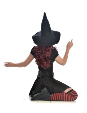 tam uzunlukta portresi kırmızı saçlı kız kara cadı kostüm ve sivri şapka giyiyor. beyaz stüdyo arka plan üzerinde izole oturmuş poz.