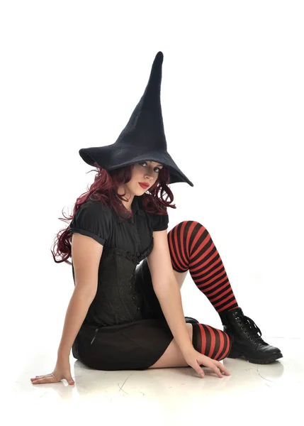 黒魔女の衣装と先のとがった帽子を身に着けている赤い髪の女の子の完全な長さの肖像画 ホワイト スタジオの背景で隔離の座りポーズ — ストック写真