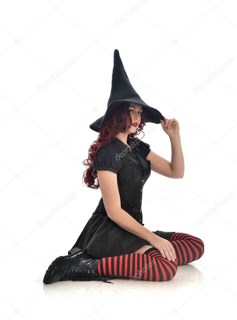 full length portrait of red haired girl wearing black witch costume and pointy hat.  seated pose, isolated on white studio background.