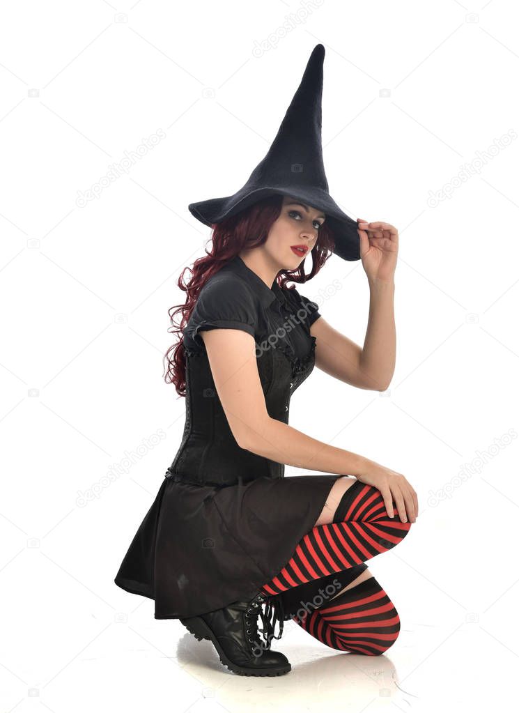 full length portrait of red haired girl wearing black witch costume and pointy hat.  seated pose, isolated on white studio background.