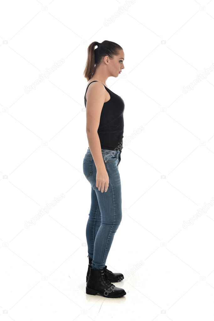 full length portrait of brunette girl wearing black single and jeans. standing pose, side profile. isolated on white studio background.