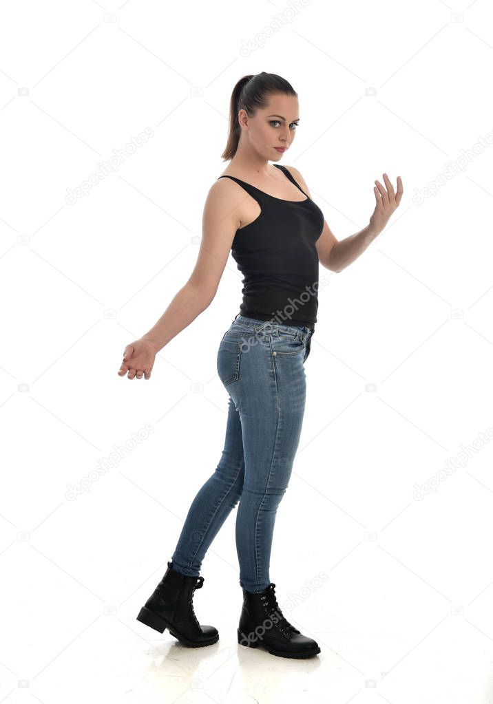 full length portrait of brunette girl wearing black single and jeans. standing pose, side profile. isolated on white studio background.