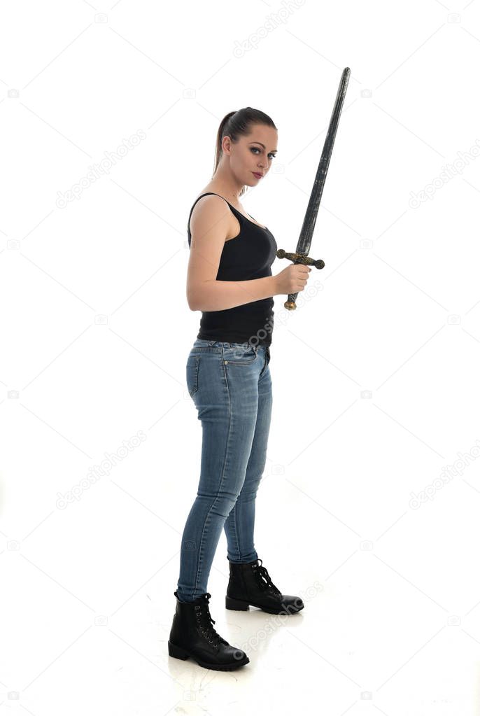 full length portrait of brunette girl wearing black single and jeans. standing pose, holding a sword. isolated on white studio background.