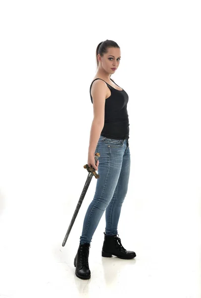 穿黑色单和牛仔裤的黑发女孩的全长肖像 站立姿势 拿着一把剑 在白色工作室背景下被隔离 — 图库照片