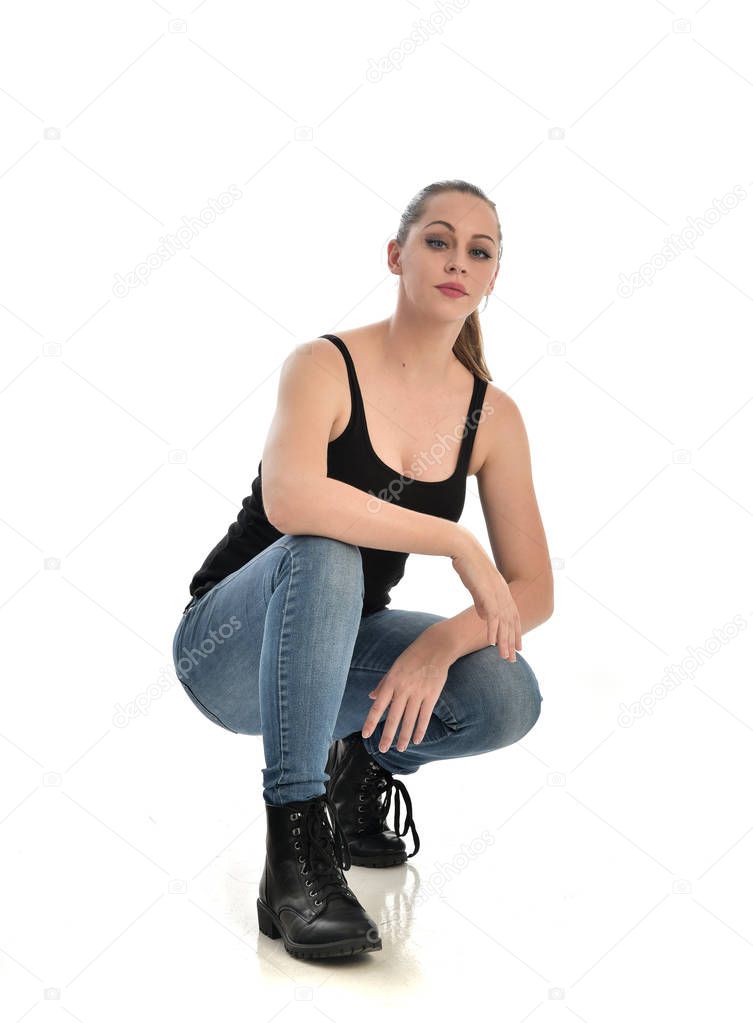 full length portrait of brunette girl wearing black single and jeans. seated pose. isolated on white studio background.