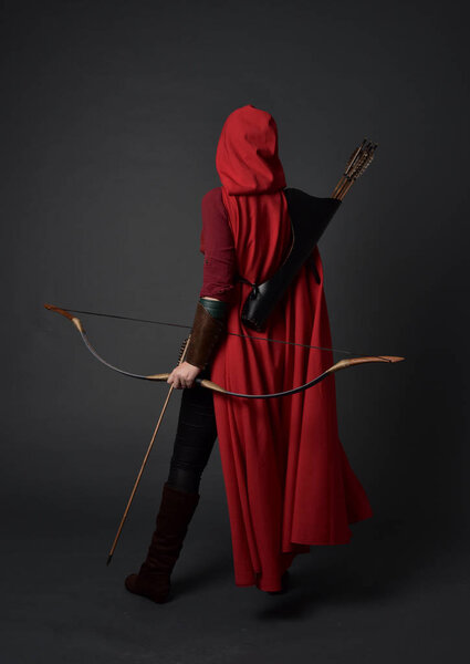 full length portrait of brunette girl wearing red medieval costume and cloak, holding a bow and arrow. standing pose on grey studio background.