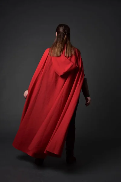 赤中世の衣装とマントを身に着けているブルネットの女の子の完全な長さの肖像画 灰色のスタジオの背景にカメラに背中と立ちポーズします — ストック写真