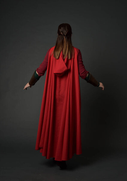 full length portrait of brunette girl wearing red medieval costume and cloak. standing pose  with back to the camera on grey studio background.