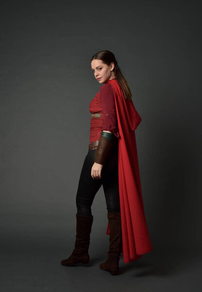 full length portrait of brunette girl wearing red medieval costume and cloak. standing pose   on grey studio background.