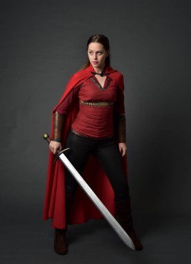 tam uzunlukta kırmızı Ortaçağ kostüm ve pelerin giymiş esmer kız portresi. gri studio arka plan üzerinde bir kılıç holding ayakta poz.