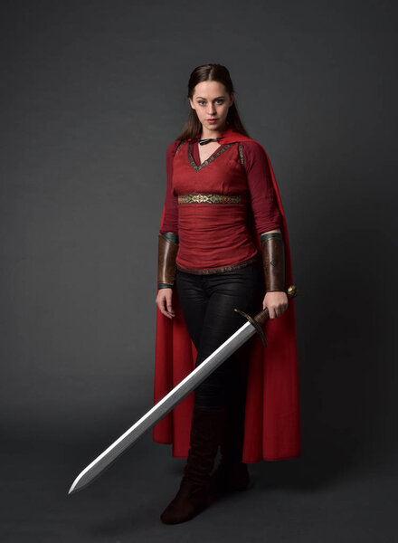 full length portrait of brunette girl wearing red medieval costume and cloak. standing pose  holding a sword on grey studio background.