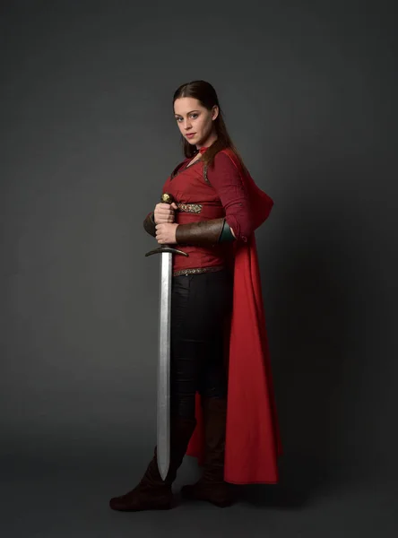 赤中世の衣装とマントを身に着けているブルネットの女の子の完全な長さの肖像画 灰色のスタジオの背景に 剣を持った立ちポーズ — ストック写真