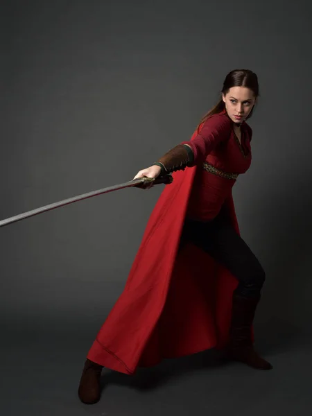 黑发女孩的全长肖像身穿红色中世纪服装和斗篷 站立姿势持有一把剑在灰色工作室背景 — 图库照片
