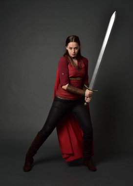 tam uzunlukta kırmızı Ortaçağ kostüm ve pelerin giymiş esmer kız portresi. gri studio arka plan üzerinde bir kılıç holding ayakta poz.