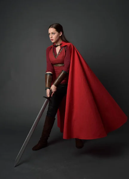 赤中世の衣装とマントを身に着けているブルネットの女の子の完全な長さの肖像画 灰色のスタジオの背景に 剣を持った立ちポーズ — ストック写真