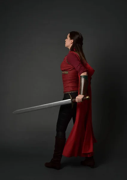 黑发女孩的全长肖像身穿红色中世纪服装和斗篷 站立姿势与回到相机 持有一把剑灰色工作室背景 — 图库照片
