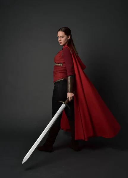 黑发女孩的全长肖像身穿红色中世纪服装和斗篷 站立姿势与回到相机 持有一把剑灰色工作室背景 — 图库照片