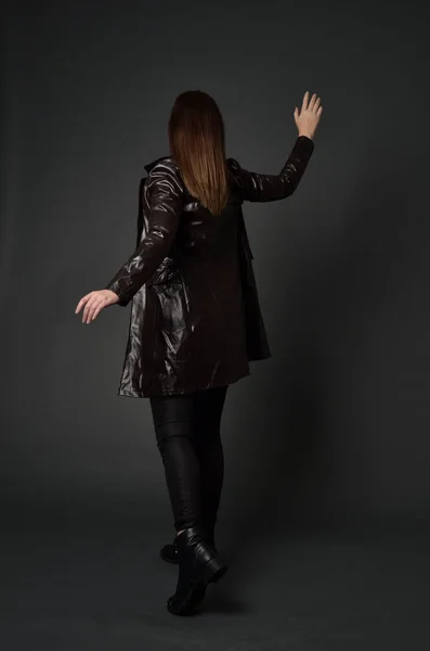 黑发女孩穿长皮革大衣和靴子全长肖像 站立姿势与回到相机 在灰色工作室背景 — 图库照片