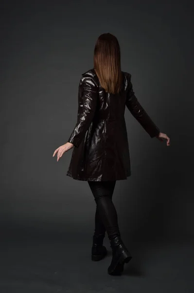 長い革のコートやブーツを身に着けているブルネットの女の子の完全な長さの肖像画 灰色のスタジオの背景に カメラに背中を立ちポーズします — ストック写真