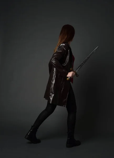 full length portrait of brunette girl wearing  red long leather jack. standing pose, holding a sword on grey studio background.
