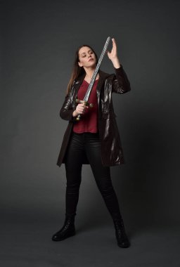 Tam boy esmer kız giyiyor kırmızı uzun deri jack portresi. poz, gri studio arka plan üzerinde bir kılıç holding ayakta.