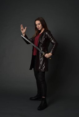 Tam boy esmer kız giyiyor kırmızı uzun deri jack portresi. poz, gri studio arka plan üzerinde bir kılıç holding ayakta.