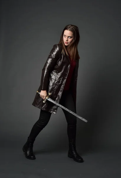 穿红色长皮革插孔的黑发女孩的全长肖像 站在姿势 拿着剑在灰色的工作室背景 — 图库照片