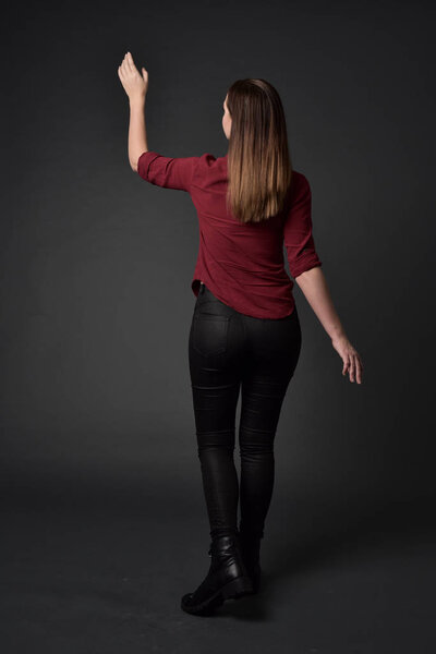 full length portrait of brunette girl wearing  red shirt and leather pants. standing pose , on grey studio background.
