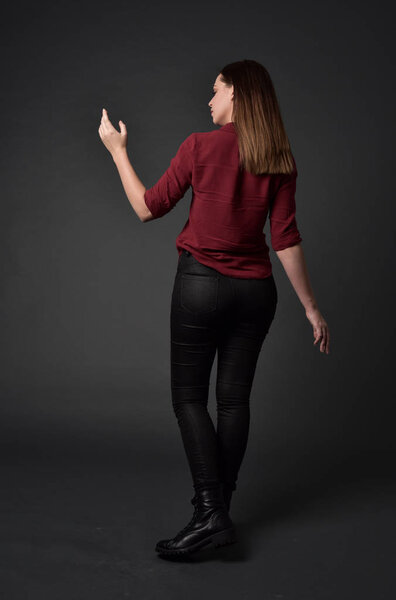full length portrait of brunette girl wearing  red shirt and leather pants. standing pose , on grey studio background.