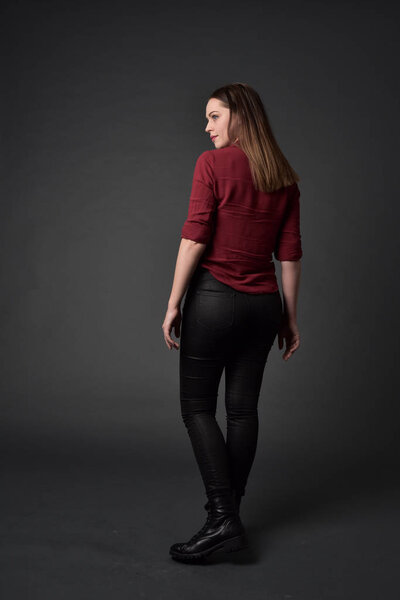 full length portrait of brunette girl wearing  red shirt and leather pants. standing pose , on grey studio background.