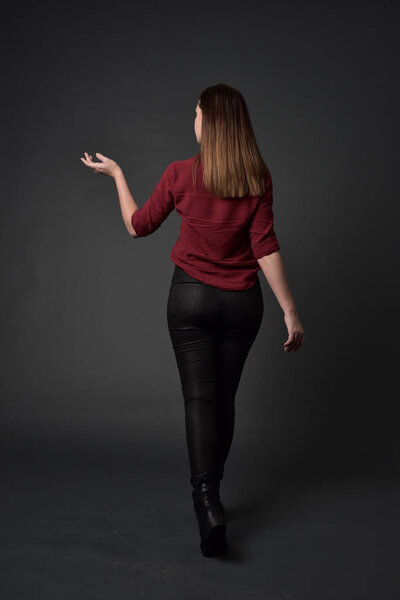 full length portrait of brunette girl wearing  red shirt and leather pants. standing pose , on grey studio background.