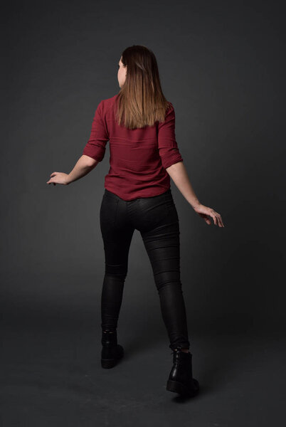 full length portrait of brunette girl wearing  red shirt and leather pants. standing pose , on grey studio background.