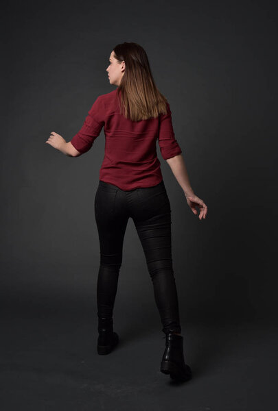 full length portrait of brunette girl wearing  red shirt and leather pants. standing pose , on grey studio background.