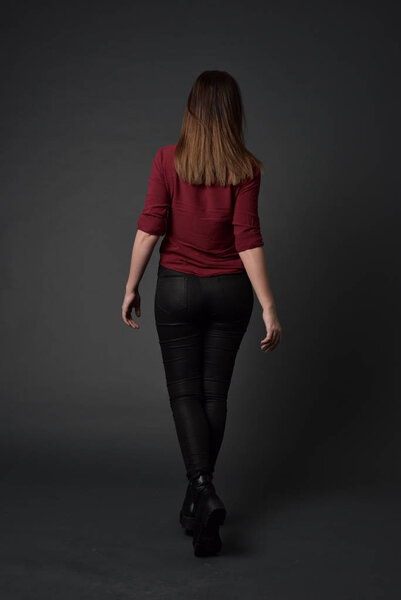 full length portrait of brunette girl wearing  red shirt and leather pants. standing pose , on grey studio background.