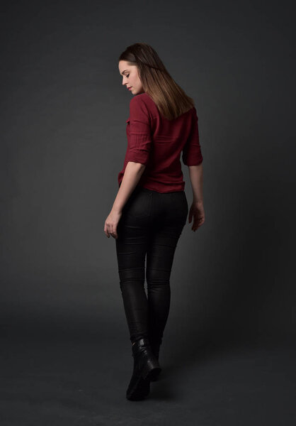 full length portrait of brunette girl wearing  red shirt and leather pants. standing pose , on grey studio background.