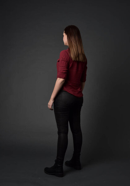 full length portrait of brunette girl wearing  red shirt and leather pants. standing pose , on grey studio background.