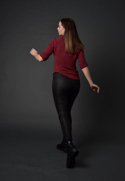 full length portrait of brunette girl wearing  red shirt and leather pants. standing pose , on grey studio background.