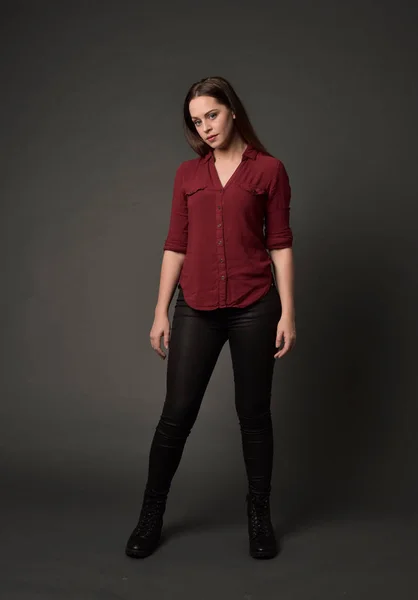 full length portrait of brunette girl wearing  red shirt and leather pants. standing pose , on grey studio background.