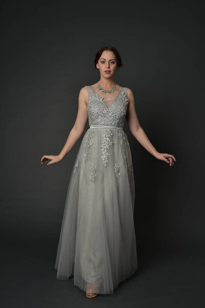 full length portrait of brunette  girl wearing long silver ball gown. standing pose on grey studio background.