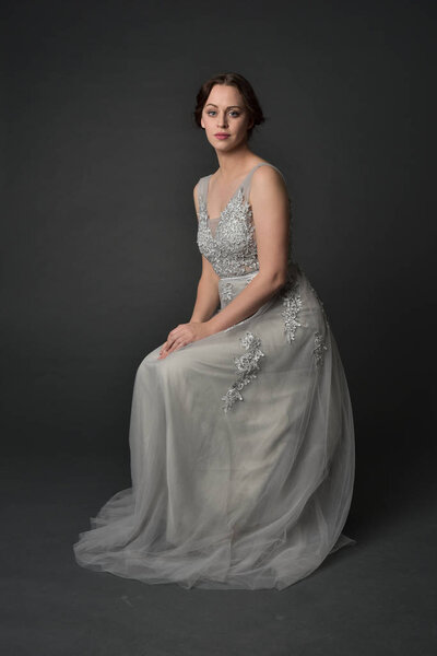 full length portrait of brunette  girl wearing long silver ball gown. seated pose on grey studio background.