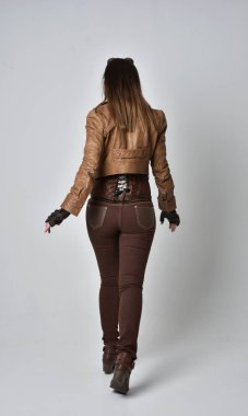 tam uzunlukta kahverengi deri steampunk kıyafeti giymiş esmer kız portresi. ayakta poz ile kameraya gri studio arka arkaya.