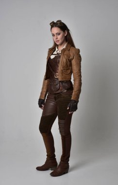 tam uzunlukta kahverengi deri steampunk kıyafeti giymiş esmer kız portresi. gri studio arka plan üzerinde ayakta poz.
