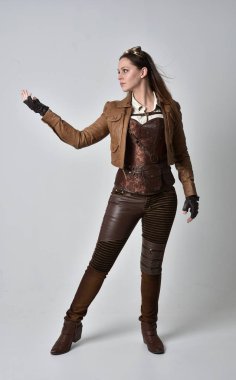 tam uzunlukta kahverengi deri steampunk kıyafeti giymiş esmer kız portresi. gri studio arka plan üzerinde ayakta poz.