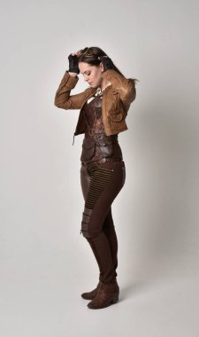 tam uzunlukta kahverengi deri steampunk kıyafeti giymiş esmer kız portresi. gri studio arka plan üzerinde ayakta poz.