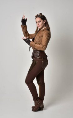 tam uzunlukta kahverengi deri steampunk kıyafeti giymiş esmer kız portresi. gri studio arka plan üzerinde ayakta poz.