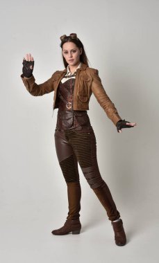 tam uzunlukta kahverengi deri steampunk kıyafeti giymiş esmer kız portresi. gri studio arka plan üzerinde ayakta poz.