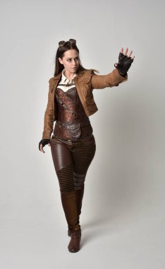 tam uzunlukta kahverengi deri steampunk kıyafeti giymiş esmer kız portresi. gri studio arka plan üzerinde ayakta poz.
