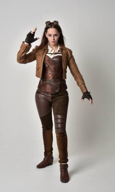 tam uzunlukta kahverengi deri steampunk kıyafeti giymiş esmer kız portresi. gri studio arka plan üzerinde ayakta poz.